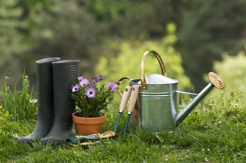In a neatly maintained outdoor garden space, a pair of black rubber boots stands on lush green grass, accompanied by a small terracotta flower pot filled with blooming purple flowers. Nearby, gardening tools including a pair of pruners, a hand trowel, and a cultivator are arranged next to a silver metal watering can with a curved handle, placed on the grass. The background features a softly blurred view of trees and foliage, suggesting a natural, landscaped setting. The scene captures a peaceful, well-tended garden area suitable for general outdoor gardening and lawn care activities, reflecting the services provided by Pressure Washing Battersea in the Battersea postcode area. The lighting indicates a clear, bright day, emphasizing the vibrant natural tones of green grass, flowering plants, and garden equipment, illustrating an environment ready for ongoing maintenance or gardening projects.